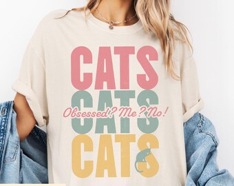 May include: Ivory-colored t-shirt with the word "CATS" stacked three times in pink, teal, and yellow. The phrase "Obsessed? Me? No!" is in the center. A small cat silhouette is at the bottom. The shirt is part of a cat-themed collection.