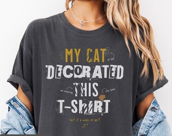 May include: Dark gray t-shirt with the text "MY CAT DECORATED THIS T-SHIRT" in white and gold lettering. The shirt has a relaxed fit and features cat-related doodles. The word "PEPPER" is at the bottom.