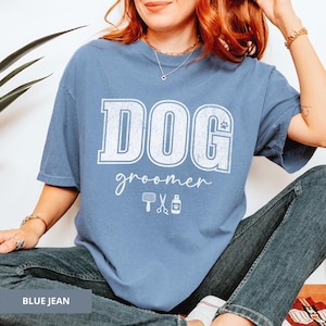 May include: A blue jean-colored t-shirt with the words "DOG groomer" in white block letters. The design includes a paw print and grooming tools. The shirt is a casual style.