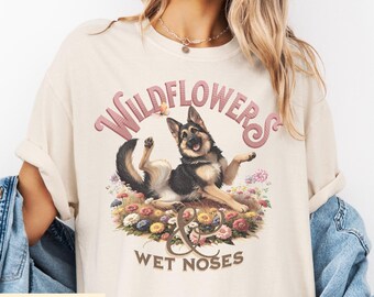 Puede incluir: Camiseta de color marfil con un gráfico de un perro pastor alemán acostado en un lecho de flores. El texto "WILDFLOWERS & WET NOSES" está arqueado sobre el perro en rosa. Un diseño de mariposa también está incluido.