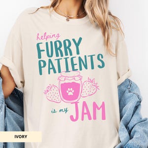 May include: Ivory-colored t-shirt with the text "helping FURRY PATIENTS is my JAM" in pink and teal. The design includes a jar of jam with a paw print and two strawberries. The shirt is a casual, oversized fit.