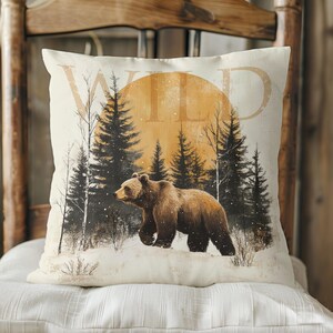 May include: A throw pillow with a brown bear walking through a snowy forest with a large orange sun in the background. The text "WILD" is printed on the pillow.