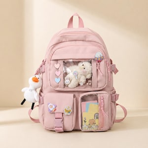 May include: A light pink backpack featuring a clear front pocket containing a white plush toy, a second clear pocket with colorful paper inserts, multiple small decorative patches, and a white duck plush keychain attached to the side strap.