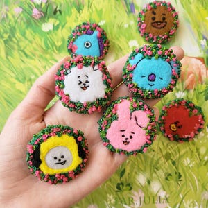 May include: A collection of colorful, felt character pins, each with a unique face and surrounded by a floral wreath. The pins feature various colors including blue, pink, yellow, and brown. The pins are handmade and have embroidered details.