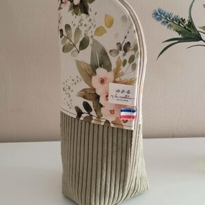 Peut inclure: Une trousse zippée verticale avec un motif floral sur la partie supérieure, composé de fleurs roses et de feuilles vertes sur fond crème. La partie inférieure est en velours côtelé vert sauge. Une petite étiquette indique les camillettes.