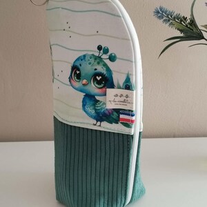 Peut inclure: Une trousse zippée verticale composée d'une partie supérieure blanche avec une illustration de paon et d'une partie inférieure en velours côtelé vert foncé. Une étiquette avec le texte "les comillettes" et un petit drapeau français.