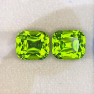 以下が含まれることがあります： 鮮やかなライムグリーンの色合いとファセットカットが施された、2つのルースクッションカットジェムストーン。角が丸みを帯びた正方形の形状で、透明な石全体に高い透明度と内部の光の反射が見られます。