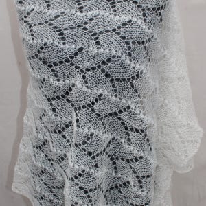 Könnte beinhalten: Ein weißer, offen gewebter Schal mit einem zarten, spitzenartigen Muster. Der Schal ist über eine Form drapiert und zeigt sein kompliziertes Design und seine weiche Textur. Die Kanten haben einen gewellten Abschluss, der zu seinem eleganten Reiz beiträgt. Ein leichtes und luftiges Accessoire.