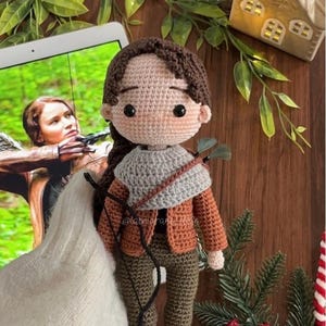 May include: A handmade crochet doll with brown hair, wearing a gray scarf, brown jacket, and green pants. The doll has black eyes and is holding a bow and arrow. The doll is on a wooden surface with a tablet showing a movie scene.