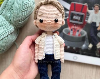 May include: A handmade crochet doll with light brown hair, wearing a cream-colored jacket, a white shirt, and navy blue pants. The doll has black eyes and white shoes. A ball of light green yarn is in the background.