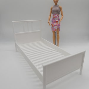 May include: White doll bed frame with slatted base and a simple design. The bed is made of plastic and is approximately 12 inches long.