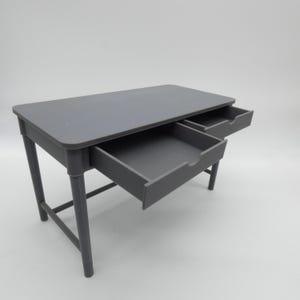 Könnte beinhalten: Ein grauer Schreibtisch mit zwei offenen Schubladen. Der Schreibtisch hat eine rechteckige Oberseite und vier abgerundete Beine. Die Schubladen sind ebenfalls grau und haben ein schlichtes, minimalistisches Design. Der Schreibtisch ist ca. 76 cm lang und 51 cm breit.