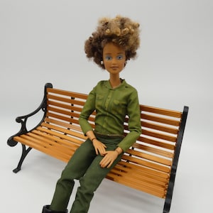 May include: A doll with brown skin and curly brown and blonde hair is seated on a wooden park bench. The doll wears a green jumpsuit and black boots. The bench has black metal supports and wooden slats.