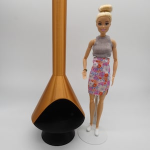May include: A gold-colored, mid-century modern style fireplace with a black interior and base. A doll wearing a gray top and floral print skirt stands next to the fireplace. The doll has blonde hair and white shoes.