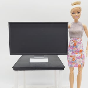 May include: A miniature black computer monitor on a black desk with white legs. A doll is standing next to the desk, wearing a grey top and a floral skirt. The monitor has a rectangular screen and a white stand.