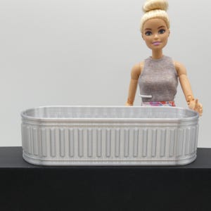 Puede incluir: Una pequeña bañera rectangular de color plateado con detalles acanalados verticales. La bañera se muestra sobre una superficie negra, con una muñeca de pie detrás. La muñeca lleva un top gris y una falda floral.