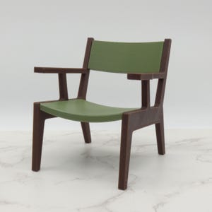 May include: A miniature armchair featuring a dark brown frame with a solid structure, including four legs and two armrests. The seat and the backrest are finished in a matte olive green color, providing a contrast to the dark brown frame.