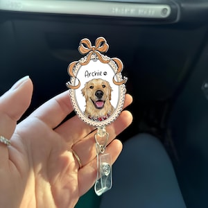 May include: A personalised badge reel with a Golden Retriever illustration, the name "Archie", and a paw print. The oval design is framed with a decorative border and a bow, with a clear plastic clip.
