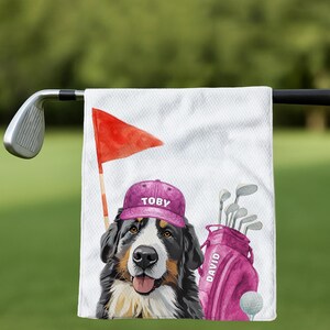 May include: A white golf towel featuring a Bernese Mountain Dog wearing a purple cap with "TOBY" printed on it. A red flag, golf bag with "DAVID" printed on it, golf clubs, and a golf ball are also visible. The background is blurred green.