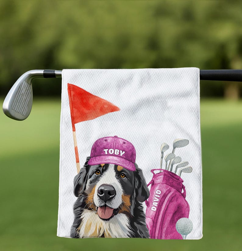 May include: A white golf towel featuring a Bernese Mountain Dog wearing a purple cap with "TOBY" printed on it. A red flag, golf bag with "DAVID" printed on it, golf clubs, and a golf ball are also visible. The background is blurred green.