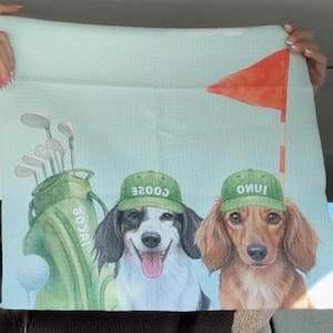 May include: A light blue golf-themed towel featuring two dogs wearing green caps with text. A golf bag, golf clubs, and a golf ball are also depicted. A red flag on a pole is in the background.