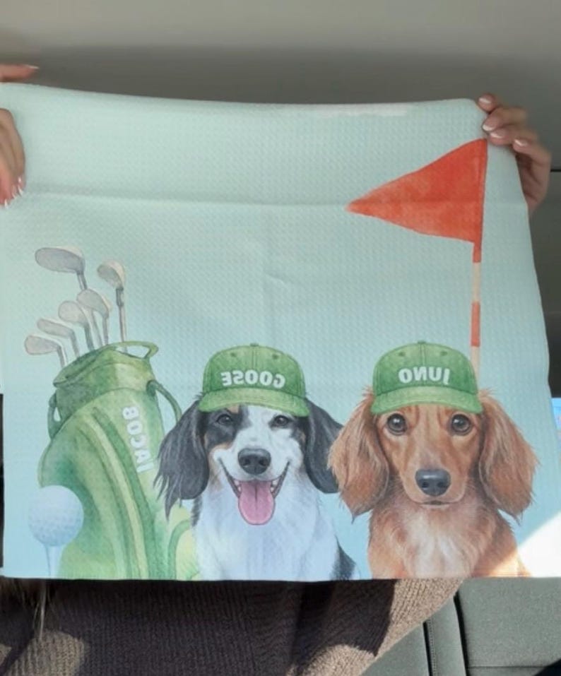 May include: A light blue golf-themed towel featuring two dogs wearing green caps with text. A golf bag, golf clubs, and a golf ball are also depicted. A red flag on a pole is in the background.