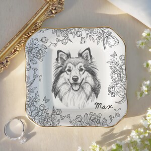 May include: A square, white ceramic trinket dish with a gold rim, featuring a black and white illustration of a dog's face. Floral and butterfly designs surround the dog. The name "Max" is written in script.