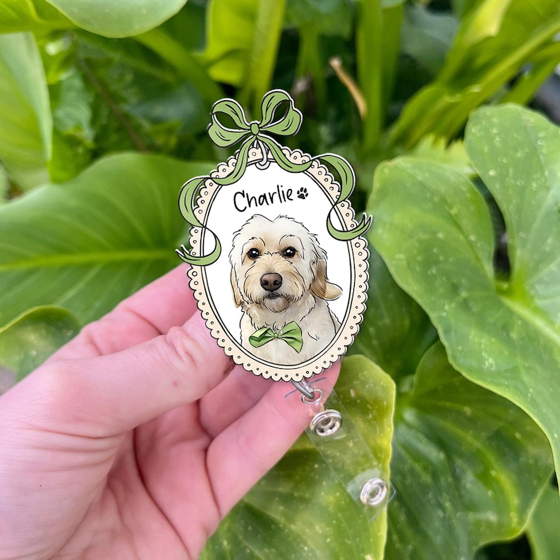 May include: An oval badge reel with a dog portrait, named Charlie, wearing a green bow tie. The design includes a green bow and ribbon. The badge reel has a retractable cord and is set against a green background.