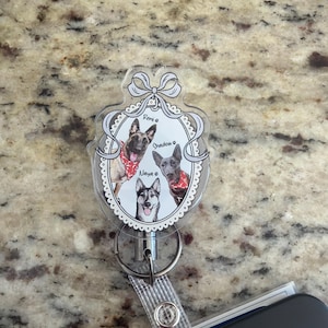 May include: A clear, oval badge reel with a decorative frame holding three dog portraits. The dogs, named Remi, Shadow, and Naya, wear red bandanas. The reel has a retractable cord and a clear strap.