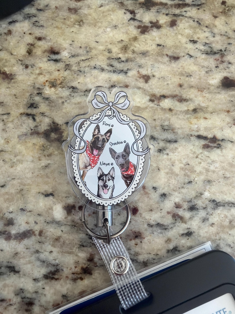 May include: A clear, oval badge reel with a decorative frame holding three dog portraits. The dogs, named Remi, Shadow, and Naya, wear red bandanas. The reel has a retractable cord and a clear strap.