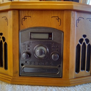 Puede incluir: Un tocadiscos de estilo vintage de madera con reproductor de CD, sintonizador de radio y reproductor de cassette. El reproductor tiene una cara plateada con botones y perillas negras. El gabinete de madera es de color marrón claro con tallas decorativas.