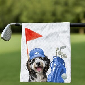 May include: White golf towel featuring a cartoon dog wearing a blue cap with "ACE" on it. A red flag, golf bag, and golf ball are also depicted. The golf bag has the name "DAVID" on it.