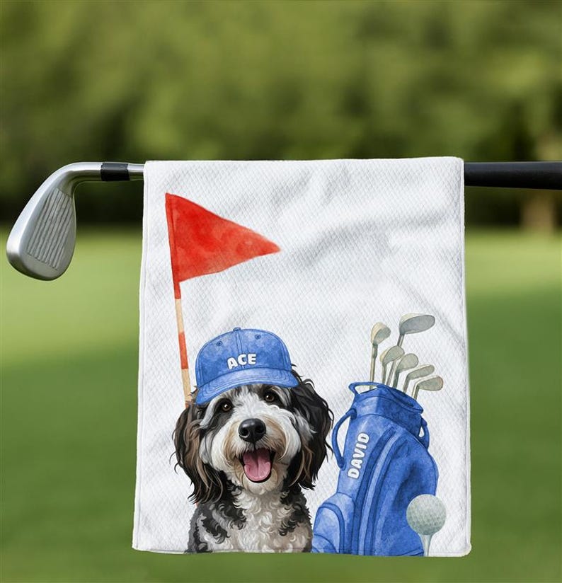 May include: White golf towel featuring a cartoon dog wearing a blue cap with "ACE" on it. A red flag, golf bag, and golf ball are also depicted. The golf bag has the name "DAVID" on it.