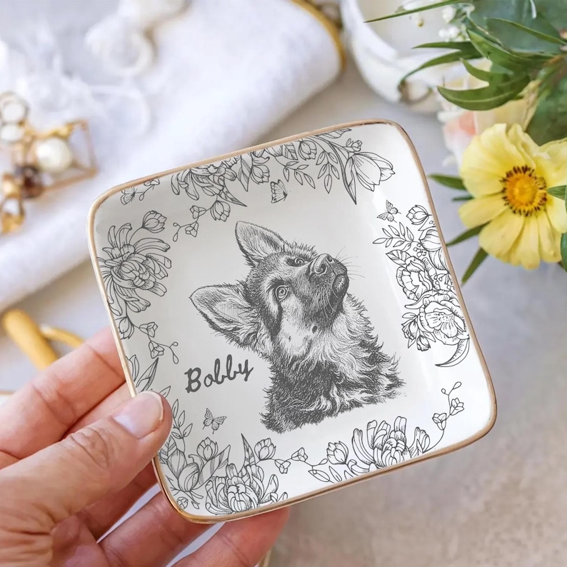 May include: Square ceramic trinket dish with a gold rim, featuring a black and white illustration of a German Shepherd dog named "Bobby." The dish is decorated with floral and butterfly designs.