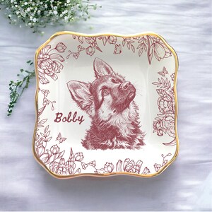 May include: Square ceramic dish with a gold rim and a maroon illustration of a German Shepherd dog named "Bobby." The dish is decorated with floral and butterfly motifs.