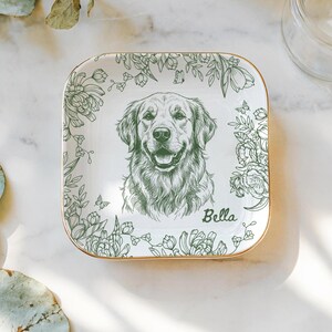 May include: Square white ceramic dish with a gold rim, featuring a detailed green sketch of a Golden Retriever dog. The dog's name, "Bella," is written in script. Floral and butterfly designs frame the dog portrait.