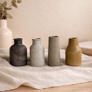 May include: Four ceramic vases in varying shapes and colors sit on a neutral-toned cloth. The vases include a dark brown ribbed vase, a light gray vase, a tall gray vase, and a mustard yellow ribbed vase.