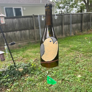 May include: A glass bottle wind chime with a painted bird design. The bottle is brown and the bird is beige with gray wings and blue eyes. A green glass piece hangs below the bottle.