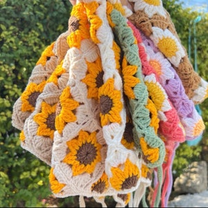 May include: A stack of crocheted sunflower-shaped bags in various colors, including white, beige, green, pink, and purple. The bags are made with a granny square pattern and have long ties.
