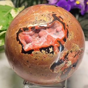 May include: A polished, spherical gemstone with a mix of brown, orange, and black hues. The sphere has a cavity revealing a cluster of pink crystals. The gemstone is displayed on a clear stand, showcasing its natural beauty.