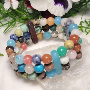 May include: A multi-strand bracelet featuring a variety of colorful gemstone beads. The bracelet includes beads in shades of blue, brown, green, orange, and pink. The bracelet is displayed on a white surface with flowers in the background.