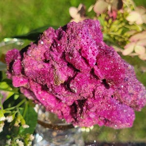 May include: A vibrant magenta-colored mineral cluster with a rough, crystalline texture. The specimen features deep pink hues and numerous small, sparkling facets across its irregular, jagged surface, reflecting light from multiple angles.