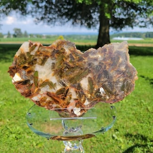 May include: A flat, irregular mineral specimen featuring a mix of translucent white quartz and embedded, elongated crystals in shades of olive green, amber, and deep brown, displayed on a clear, circular glass stand with a faceted pedestal base.