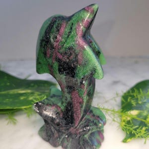May include: A carved stone dolphin figurine. The sculpture features a green, black, and magenta color scheme. The dolphin is depicted leaping, with its tail and base integrated into the stone. The figurine is set against a blurred background.