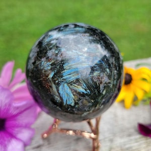 May include: A polished, spherical larvikite stone featuring a dark gray and black base with iridescent blue flashes throughout the surface. The sphere is displayed on a thin, copper-colored metal tripod stand with a branch-like design.