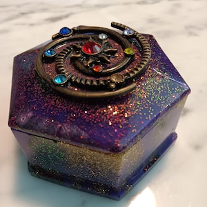 Puede incluir: Una caja hexagonal de resina púrpura con purpurina dorada y una tapa de bronce con temática de galaxia con diamantes de imitación de colores.