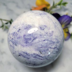 May include: A polished sphere with a marbled appearance, featuring a blend of white and lavender hues. The spherical object has a smooth surface with intricate veining patterns. The background includes colorful flowers.