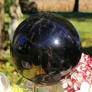 May include: A polished, dark purple fluorite sphere with visible internal fractures and natural inclusions, resting on a clear acrylic stand. The surface of the stone reflects light, showing deep violet tones and subtle crystalline patterns throughout the orb.