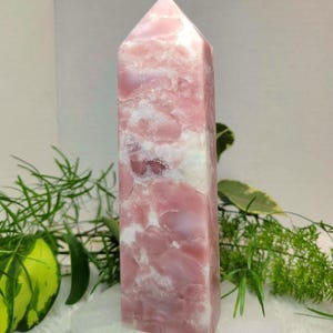 May include: A pink and white crystal obelisk. The polished stone has a rectangular base and a pointed top. The pink colour is swirled with white, creating a marbled effect. The crystal is displayed on a white surface with green foliage in the background.