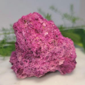May include: A vibrant, textured mineral specimen in shades of magenta and fuchsia. The rough, crystalline surface of the rock is detailed, with variations in color and texture. The mineral is a striking example of natural formations.